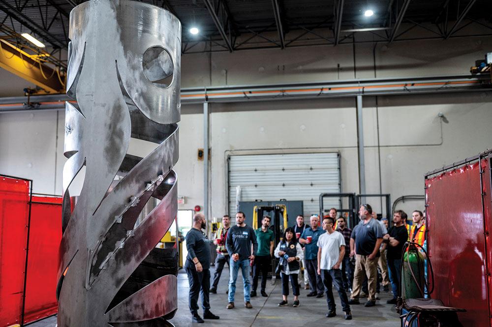 In the background, people are shown on a tour, looking at a totem designed by local artist James Harry in the foreground.