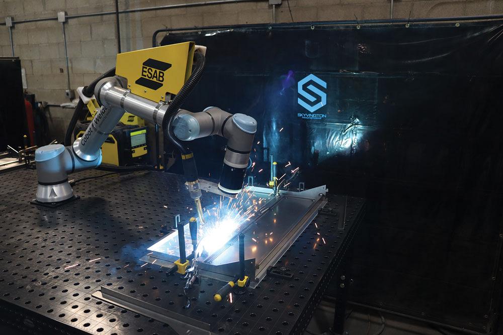 A cobot is seen mid-weld on a table-mounted assembly. 