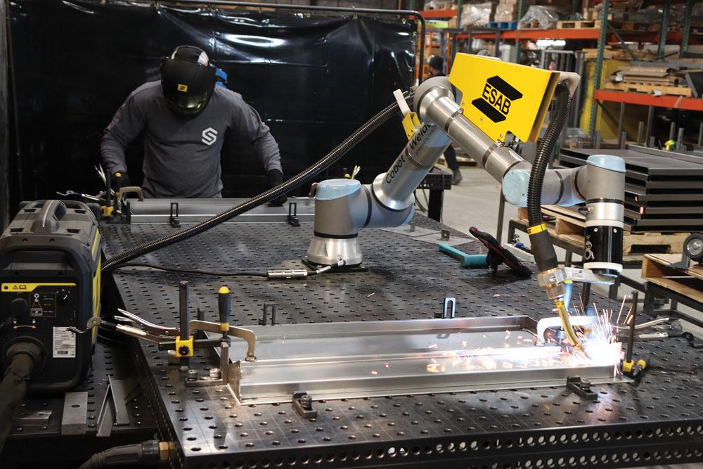 A cobot is seen completing a weld while in the background an employee prepares another assembly on the same table.