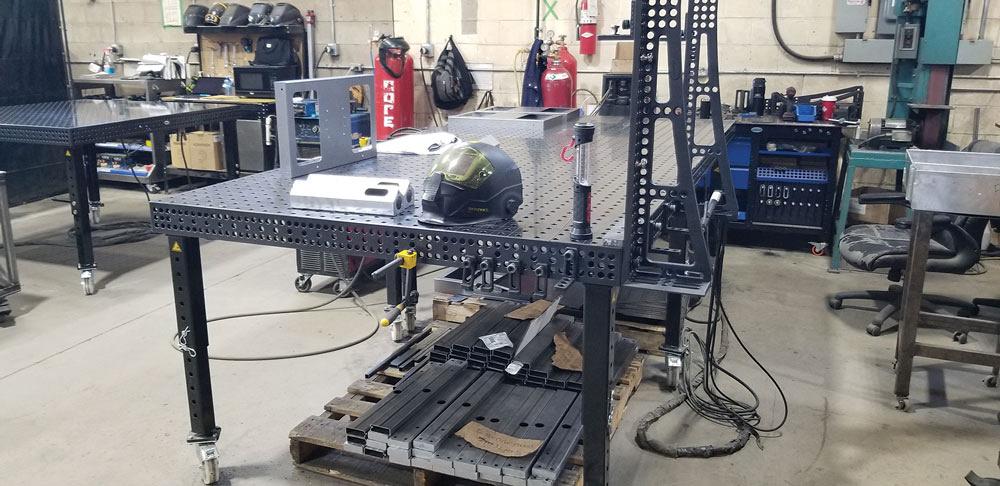 A fixture table is seen in the shop with a welding helmet rest on top of it. 