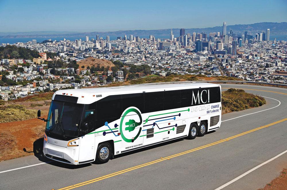 The MCI electric bus drives up a hill away from a large agglomeration.