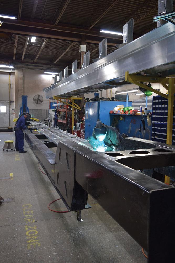 Two people wearing protective welding gear weld the base of a trailer.