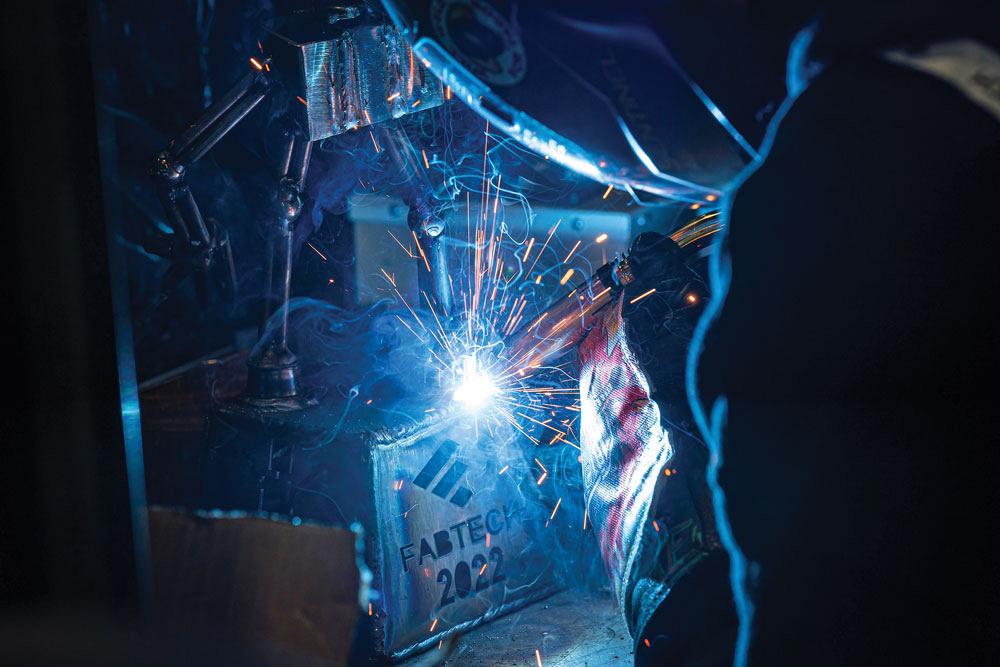 A welder is shown welding a metal box with the FABTECH logo emblazoned upon it. 