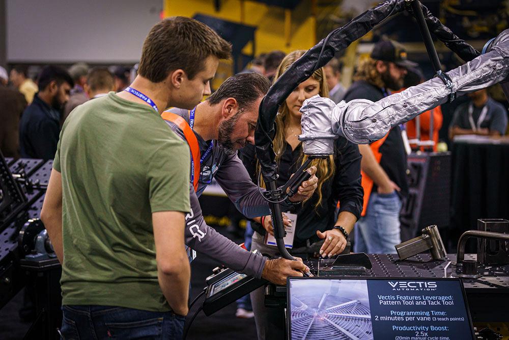 A person manipulates a welding head attached to a cobot as two show attendees observe.