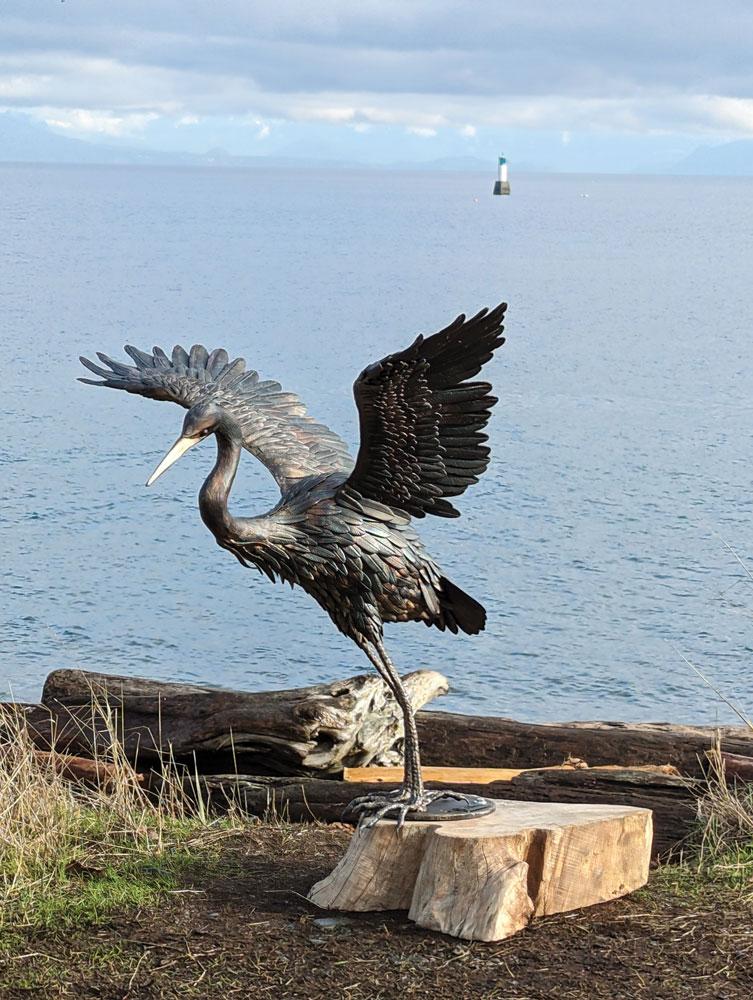The metal sculpture “Blue Heron” by Angellos Glaros stands by a body of water.  