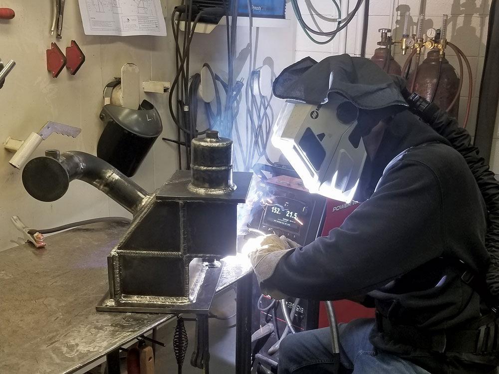 Ben Rainforth, wearing a welding helmet connected to a PAPR unit, performs a MIG weld on an assembly that sits on a work bench.  